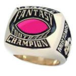 A gold ring with the words "Fantasy Football Champion" and "Mustangs" engraved on it, featuring a pink oval gemstone in the center.