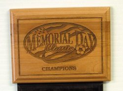 A wooden plaque with an engraved design reading "Memorial Day Classic Champions" featuring a small American flag and a soccer ball.